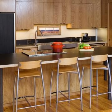 Photo of modern kitchen with wooden paneling. Island bench with 3 stools and black bench top