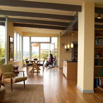 A photo of a modern home with open plan living looking from the lounge room to the dining area and kitchen. There is a young women in a manual wheelchair in the dining room.