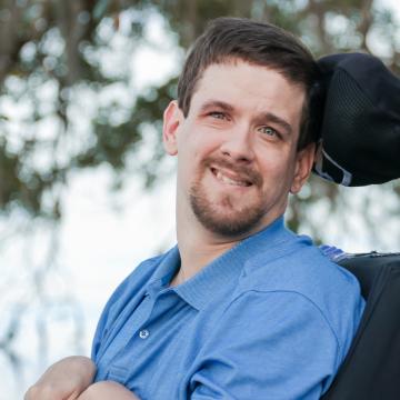 Photo of David Stoner, wearing a blue shirt, sitting in his black wheel chair with a tree in the background.