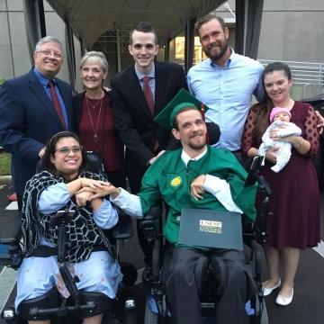 Photograph of a young woman and young man wearing a graduation cap and gown in wheelchairs holding hands with five individuals standing up behind them. One individual is holding an infant.
