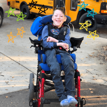 Boy with brown hair wearing a blue shirt and blue pants in a red wheelchair smiling in front of a school bus