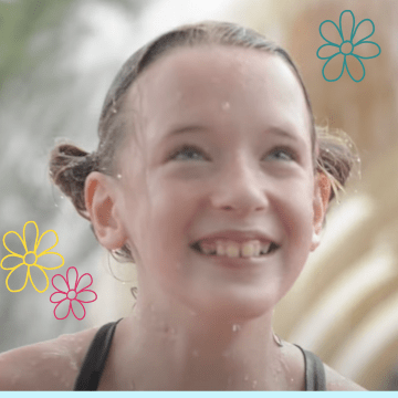 A little girl with blond pigtails and a big smile is in front of a fountain at a water park wearing her swim suit.